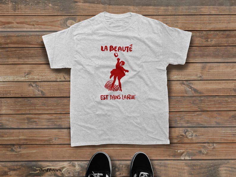 May include: A light gray short-sleeved t-shirt featuring a red graphic print of a person jumping above a net. The text "LA BEAUT&Eacute;" is printed in red above the figure, and "EST DANS LARUE" is printed in red below the figure on the chest.