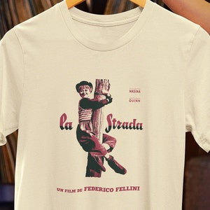May include: A beige t-shirt with a vintage-style print of a young boy climbing a pole. The print includes the text "la Strada" and "UN FILM DE FEDERICO FELLINI".