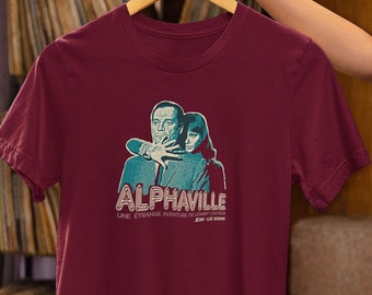 May include: A maroon t-shirt with a graphic print of a man and woman from the movie Alphaville. The text "Alphaville" is in a retro font, and the text "Une etrange aventure de Lemmy Caution" is below it.
