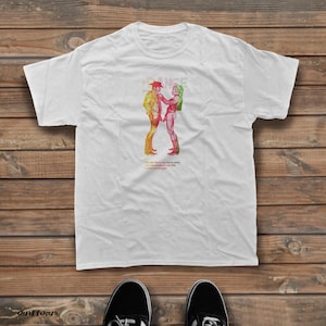 May include: A white short-sleeved t-shirt featuring a colourful graphic of two figures facing each other. The graphic includes the text DANCE at the top and smaller text below the figures. The shirt has a crew neckline and a standard fit.