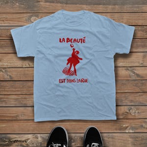 May include: A light blue short-sleeve t-shirt featuring a red graphic of a person jumping above a net. The text "LA BEAUT&Eacute;" is printed in red above the figure, and the text "EST DANS LARUE" is printed in red below the figure.