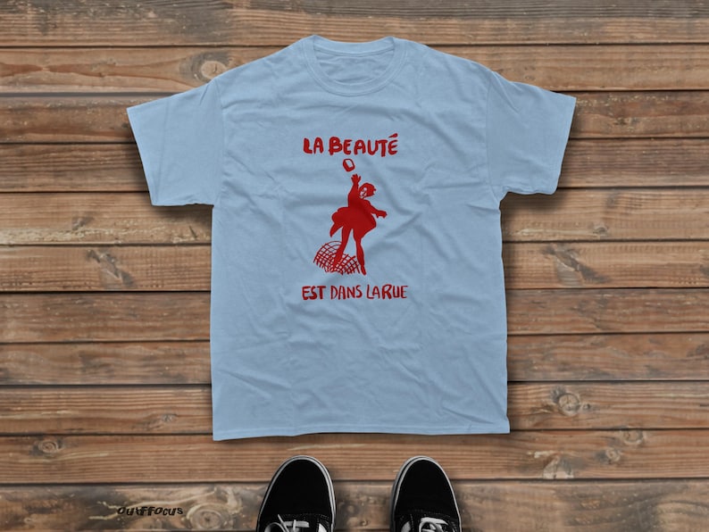 May include: A light blue short-sleeve t-shirt featuring a red graphic of a person jumping above a net. The text "LA BEAUT&Eacute;" is printed in red above the figure, and the text "EST DANS LARUE" is printed in red below the figure.
