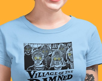 May include: A light blue t-shirt featuring a black and white graphic print of children with glowing yellow eyes. Below the illustration, the text reads Village of the Damned in a bold, stylized black font. The shirt has a classic crew neckline.