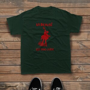 May include: A dark green short-sleeved t-shirt featuring a red graphic of a figure dancing above a grid pattern. The text "LA BEAUT&Eacute;" is printed in red above the figure, and "EST DANS LA RUE" is printed in red below the figure.