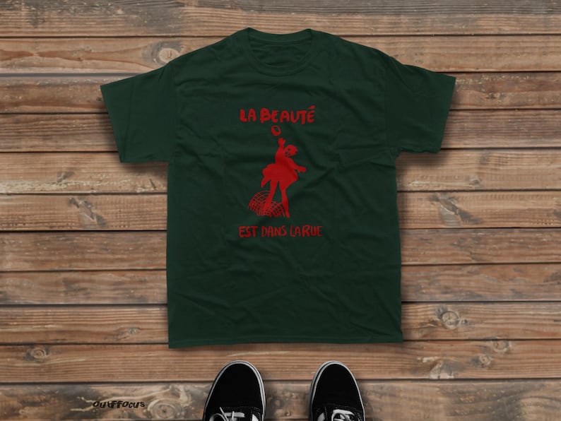 May include: A dark green short-sleeved t-shirt featuring a red graphic of a figure dancing above a grid pattern. The text "LA BEAUT&Eacute;" is printed in red above the figure, and "EST DANS LA RUE" is printed in red below the figure.