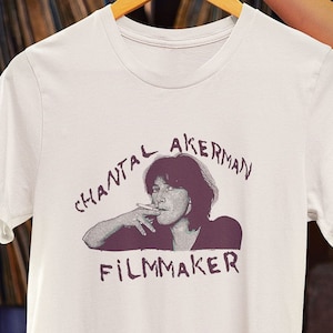 Puede incluir: Camiseta blanca con una impresión morada de Chantal Akerman, una cineasta, fumando un cigarrillo. El texto "Filmmaker" está impreso debajo de su imagen.