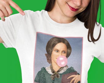 May include: A white t-shirt featuring a square graphic print of Charlotte Brontë wearing a dark green dress with a white lace collar and a dark neck scarf, blowing a large pink bubblegum bubble, with the text Charlotte Brontë in pink below.