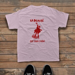 May include: A light pink short-sleeved t-shirt featuring a red graphic of a person throwing a bouquet of flowers above a curved grid pattern. The text LA BEAUT&Eacute; is printed above the graphic and EST DANS LA RUE is printed below the graphic.