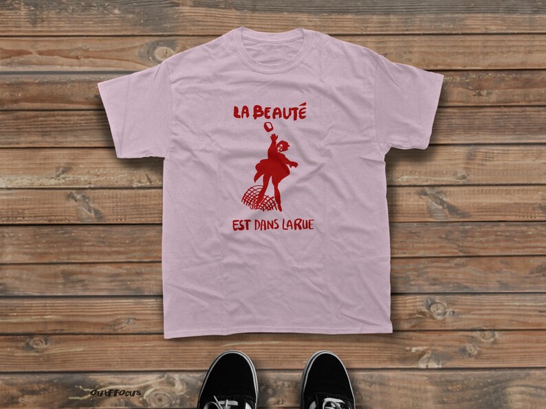 May include: A light pink short-sleeved t-shirt featuring a red graphic of a person throwing a bouquet of flowers above a curved grid pattern. The text LA BEAUT&Eacute; is printed above the graphic and EST DANS LA RUE is printed below the graphic.