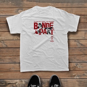 May include: A white short-sleeved t-shirt featuring a red and black graphic print of the film Bande à Part. The design includes the text Un Film De Jean-Luc Godard, the title Bande à Part, and the names Anna Karina, Sami Frey, and Claude Brasseur.