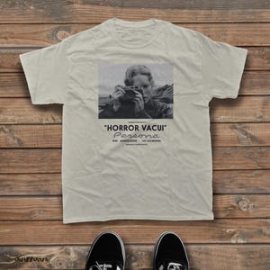 Puede incluir: Una camiseta beige de manga corta con una impresión fotográfica en blanco y negro de una persona sosteniendo una cámara. Debajo de la imagen, el texto dice: "HORROR VACUI" PERSONA, BIBI ANDERSSON, LIV ULLMANN, INGMAR BERGMAN'S.