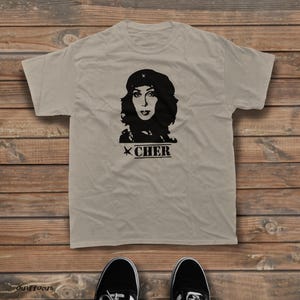 Może przedstawiać: Beżowy t-shirt z krótkim rękawem z czarnym nadrukiem graficznym przedstawiającym osobę w berecie. Pod portretem znajduje się napis CHER wydrukowany pogrubioną, stylizowaną czcionką z małą ikoną gwiazdki po lewej stronie nazwy.