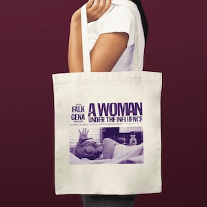 Può includere: Una borsa di tela bianca con una stampa viola del poster del film "Una donna sotto l'influenza" con Peter Falk e Gena Rowlands. Il testo sulla borsa recita "Una donna sotto l'influenza" scritto e diretto da John Cassavetes.
