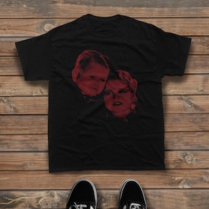 May include: A black short-sleeved t-shirt featuring a red graphic print of two faces positioned side-by-side in the center of the chest. The shirt has a crew neckline and a standard fit, displayed flat against a horizontal wooden plank background.