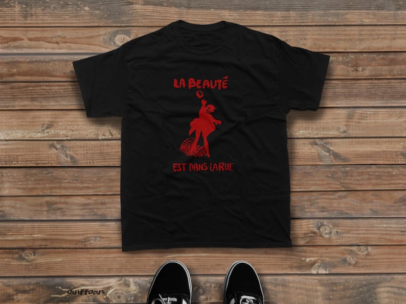 May include: A black short-sleeved t-shirt featuring a red graphic print of a person jumping above a grid pattern. The text "LA BEAUT&Eacute;" is printed in red above the figure, and "EST DANS LA RUE" is printed in red below the figure.