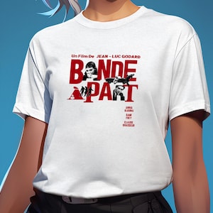 Pode incluir: Uma t-shirt branca de manga curta com uma impressão gráfica vermelha com o texto "Un Film De Jean-Luc Godard" e "Bande à Part". O design inclui fotografias a preto e branco de atores e os nomes Anna Karina, Sami Frey e Claude Brasseur.