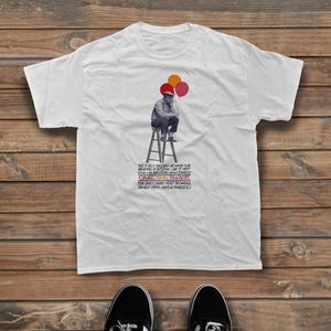 May include: A white short-sleeve t-shirt featuring a black and white graphic of a person sitting on a stool holding three balloons in orange, red, and pink. Below the graphic is text that reads: THIS IS BILLY WILDER. HE MADE THE APARTMENT & SOME LIKE IT HOT. NOW HIS EXPLOSIVE NEW COMEDY ONE, TWO, THREE STARRING JAMES CAGNEY, HORST BUCHHOLZ, PAMELA TIFFIN, ARLENE FRANCIS.