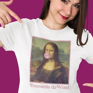 Może przedstawiać: Biała koszulka z nadrukiem obrazu Mona Lisy. Mona Lisa ma różową bańkę. Pod obrazem znajduje się napis "Leonardo da Vinci".