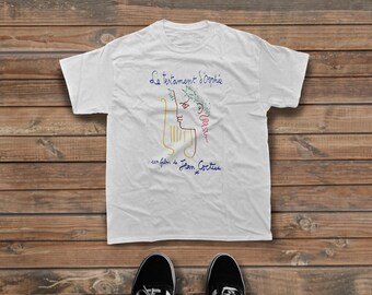 Może przedstawiać: Biały t-shirt z krótkim rękawem z kolorowym rysunkiem liniowym profilu z wieńcem laurowym i lirą. Tekst brzmi Le testament d'Orphée oraz un film de Jean Cocteau niebieską czcionką. Koszulka jest wyeksponowana na drewnianej powierzchni.