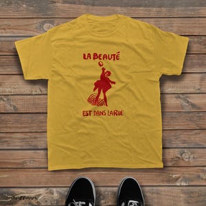 May include: A mustard yellow short-sleeve t-shirt featuring a red graphic of a person jumping over a net. The text LA BEAUT&Eacute; is printed above the figure, and the text EST DANS LA RUE is printed below the figure in a bold red font.