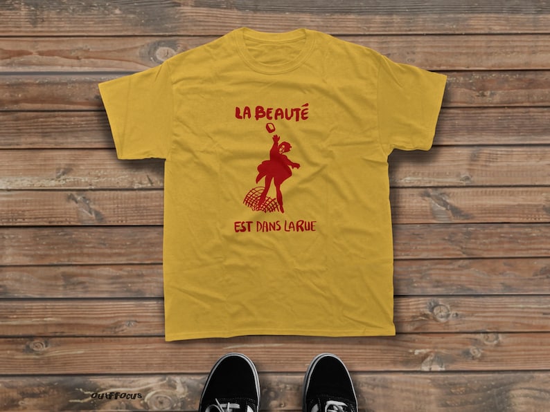 May include: A mustard yellow short-sleeve t-shirt featuring a red graphic of a person jumping over a net. The text LA BEAUT&Eacute; is printed above the figure, and the text EST DANS LA RUE is printed below the figure in a bold red font.