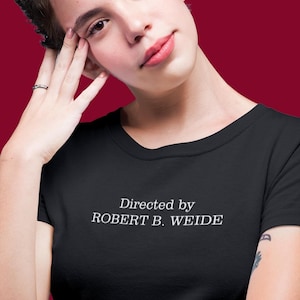 以下が含まれることがあります： 胸元にセリフ体の白い文字で「Directed by ROBERT B. WEIDE」とプリントされた、黒の半袖Tシャツ。丸首のネックラインと標準的なフィット感で、シンプルで清潔感のある日常着として着用できるデザインです。