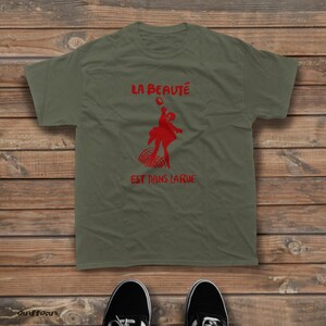 May include: An olive green short-sleeved t-shirt featuring a red graphic of a person jumping above a grid pattern. The text "LA BEAUT&Eacute;" is printed in red above the graphic, and the text "EST DANS LA RUE" is printed in red below the graphic.