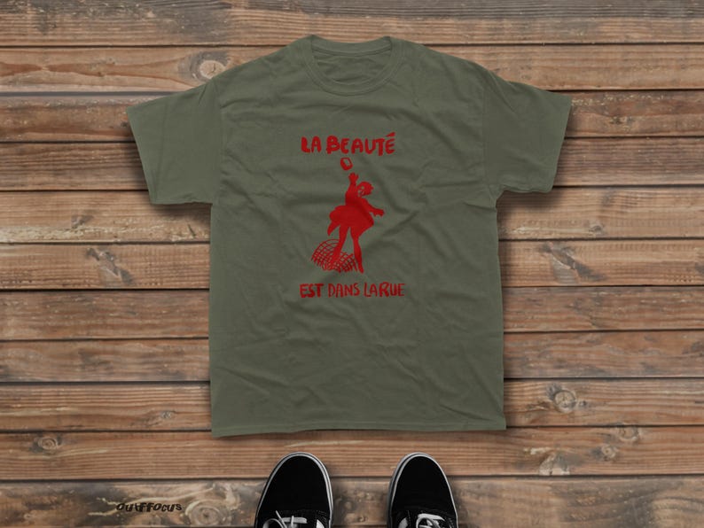 May include: An olive green short-sleeved t-shirt featuring a red graphic of a person jumping above a grid pattern. The text "LA BEAUT&Eacute;" is printed in red above the graphic, and the text "EST DANS LA RUE" is printed in red below the graphic.
