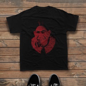 May include: A black short-sleeved t-shirt featuring a red graphic print of a smiling person with a distinctive hairstyle and a collared garment. The design uses a halftone dot pattern to create shading and detail on the central portrait.