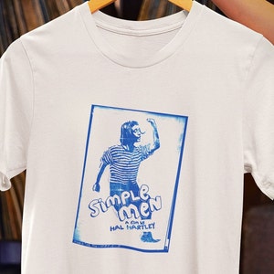 Puede incluir: Camiseta blanca con una impresión azul de un cartel de película para "Simple Men, una película de Hal Hartley". El cartel presenta a una persona con una camisa a rayas levantando el puño.