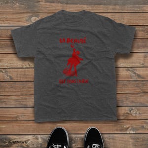 May include: A charcoal gray short-sleeve t-shirt featuring a red graphic of a person dancing and the French text LA BEAUT&Eacute; EST DANS LA RUE printed in red. The shirt has a crew neckline and a standard fit, showcasing a casual and simple design.
