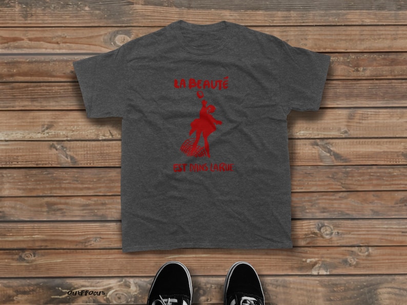 May include: A charcoal gray short-sleeve t-shirt featuring a red graphic of a person dancing and the French text LA BEAUT&Eacute; EST DANS LA RUE printed in red. The shirt has a crew neckline and a standard fit, showcasing a casual and simple design.