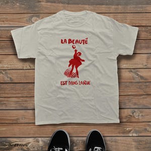 May include: A light beige short-sleeved t-shirt featuring a red graphic print of a person throwing an object above a net. The text "LA BEAUT&Eacute;" appears above the figure, and "EST DANS LARUE" is printed below the graphic in red lettering.