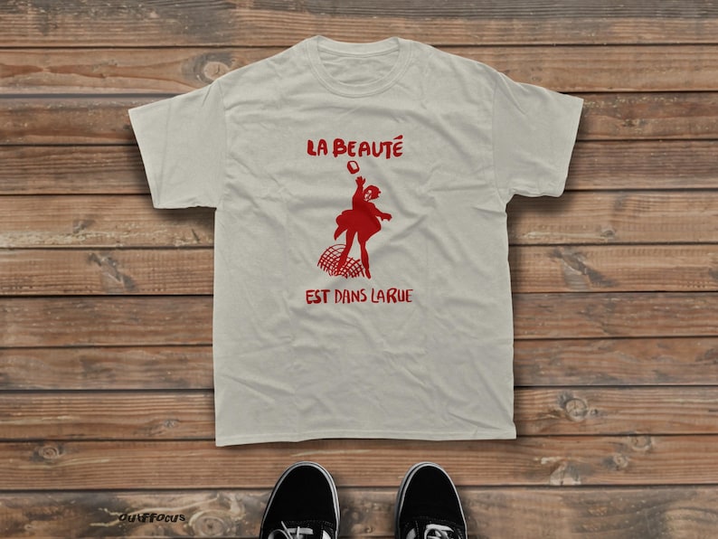 May include: A light beige short-sleeved t-shirt featuring a red graphic print of a person throwing an object above a net. The text "LA BEAUT&Eacute;" appears above the figure, and "EST DANS LARUE" is printed below the graphic in red lettering.