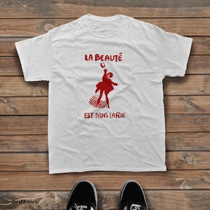 May include: A white short-sleeved t-shirt featuring a red graphic of a person throwing an object above a net. The text LA BEAUT&Eacute; EST DANS LA RUE is printed in red above and below the graphic. The shirt has a crew neckline and short sleeves.