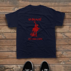 May include: A navy blue short-sleeved t-shirt featuring a red graphic of a person dancing above a curved grid pattern. The text "LA BEAUT&Eacute;" is printed in red above the graphic, and "EST DANS LA RUE" is printed in red below the graphic.