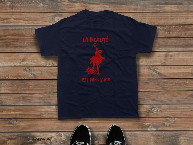 May include: A navy blue short-sleeved t-shirt featuring a red graphic of a person dancing above a curved grid pattern. The text "LA BEAUT&Eacute;" is printed in red above the graphic, and "EST DANS LA RUE" is printed in red below the graphic.