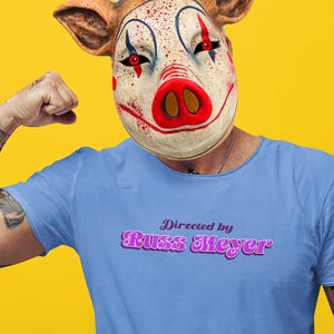 May include: A light blue t-shirt with the text "Directed by Russ Meyer" in a purple, retro font. The person is wearing a pig-clown mask with red eyes and blood splatter details. The background is yellow.