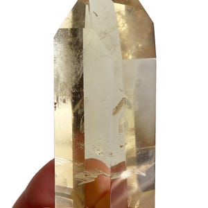 Puede incluir: Un cristal claro y facetado con un tono amarillo pálido. El cristal se sostiene en una mano y tiene una parte superior puntiaguda. La superficie del cristal tiene algunas imperfecciones e inclusiones. El cristal es un espécimen mineral natural.