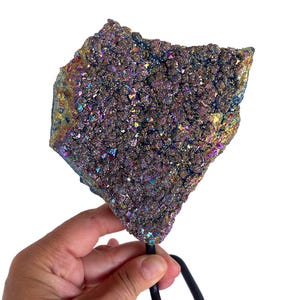 Könnte beinhalten: Große, schillernde Geodenprobe mit rauer, strukturierter Oberfläche. Die Kristallformation zeigt ein Farbspektrum, darunter Lila, Blau, Gold und Grün. Die Geode ist auf einem schwarzen Metallständer zur Präsentation montiert.