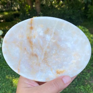Può includere: Un piatto in pietra di forma ovale con una superficie liscia e lucida, una base di colore bianco traslucido e venature minerali naturali color ambra e marrone dorato che attraversano tutto il materiale, creando un motivo unico sull'oggetto.
