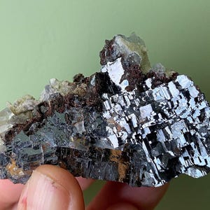 May include: A close-up of a mineral specimen featuring a cluster of metallic, silver-colored crystals with a geometric structure. The crystals are set against a backdrop of brown and beige minerals, with a light green background.