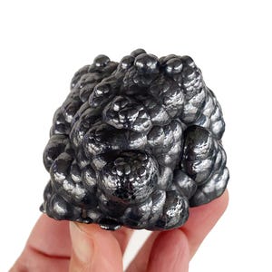 Puede incluir: Un espécimen mineral oscuro y metálico con una superficie texturizada y burbujeante. El color es un gris profundo y reflectante, con una forma ligeramente irregular. El mineral parece estar sostenido por una persona.