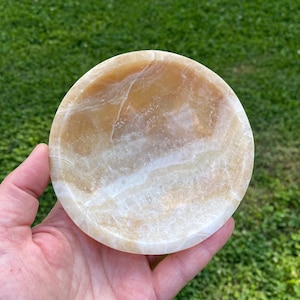 May include: A circular bowl carved from polished honey-colored onyx stone, featuring natural translucent white and golden-brown veining throughout the smooth, concave surface, showcasing the unique mineral patterns and polished texture of the material.