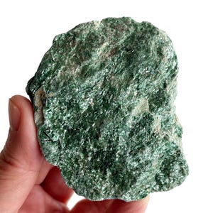 Può includere: Una mano tiene in mano un pezzo di minerale verde grezzo e di forma irregolare. Il minerale ha una superficie strutturata con inclusioni cristalline scintillanti. Il colore è un verde intenso e vibrante con macchie verdi e grigie più chiare.