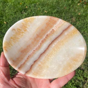 Può includere: Un piatto in pietra di forma ovale con una superficie lucida caratterizzata da motivi naturali a fasce nei toni del crema, del bianco e dell'ambra calda. La pietra presenta qualità traslucide e bordi lisci e arrotondati, mettendo in mostra le variazioni minerali.