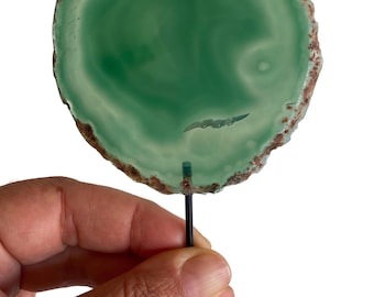 May include: A polished, circular, green agate slice with a brown, rough edge, mounted on a black metal stand. The agate features natural patterns and variations in color. The stand has a square base and a thin, vertical support.