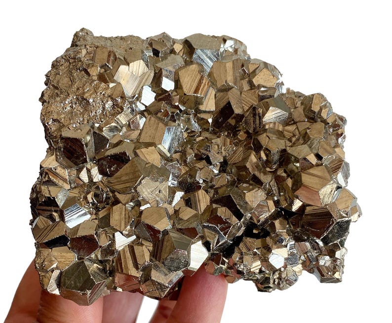May include: A cluster of pyrite crystals, also known as fool's gold, with a metallic gold color and a geometric, cubic structure. The crystals are tightly packed together, creating a textured surface. The mineral specimen is held in a hand.