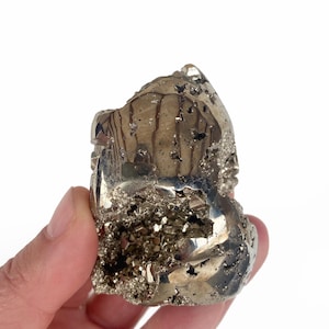 May include: A raw pyrite specimen, also known as fool's gold, held against a white background. The metallic mineral has a reflective, golden-silver lustre with a rough, textured surface. The pyrite is irregularly shaped with crystalline formations.