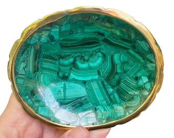 May include: An oval-shaped decorative bowl featuring a mosaic of green malachite stone fragments with distinct banded patterns. The bowl is encased in a smooth, polished gold-toned metal rim that follows the irregular, organic edge.